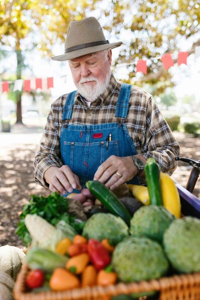 Bearded man in denim overalls standing at farmer's market with fresh vegetables, outdoors.